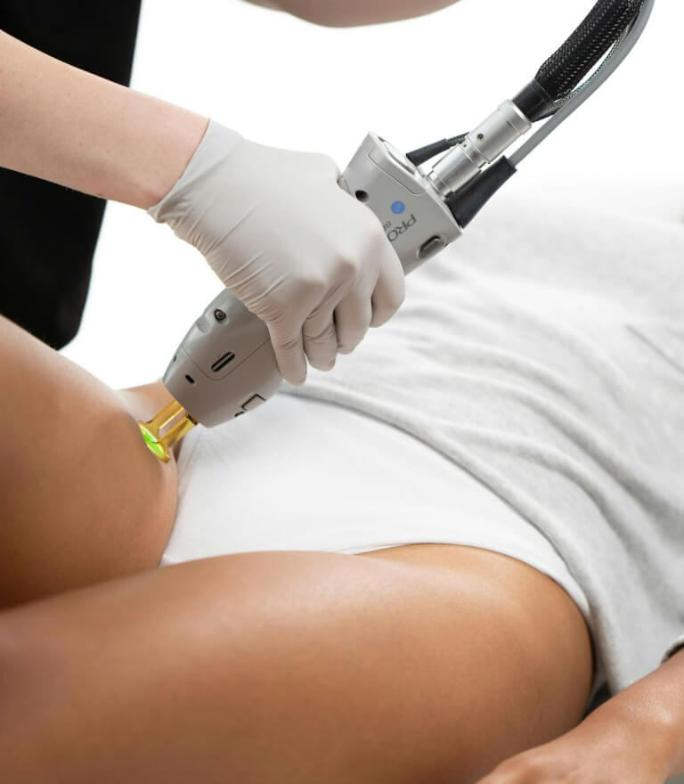 A woman receiving Laser Hair Removal treatment on her legs by a professional wearing protective eyewear and gloves in a modern clinic setting.